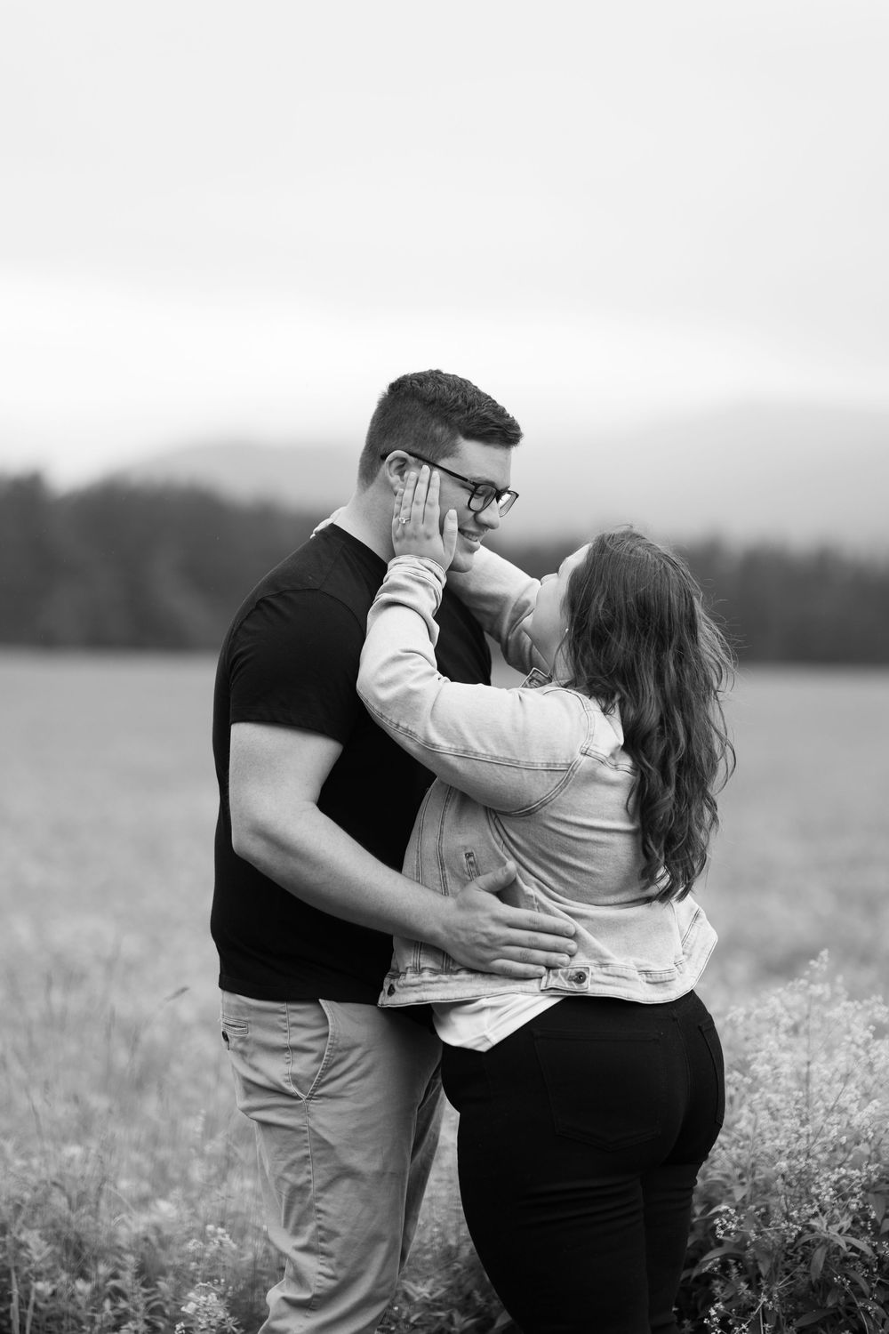 A romantic couple embraces in a field with mountains in the background, captured in black and white.