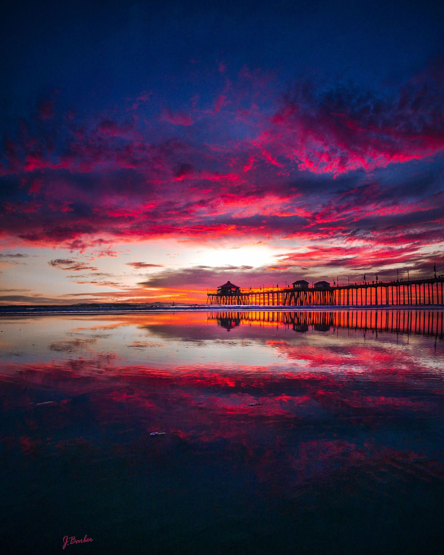 Vibrant red and purple sunset clouds reflect dramatically in still water, creating a mesmerizing mirror-like panorama at hungtington beach