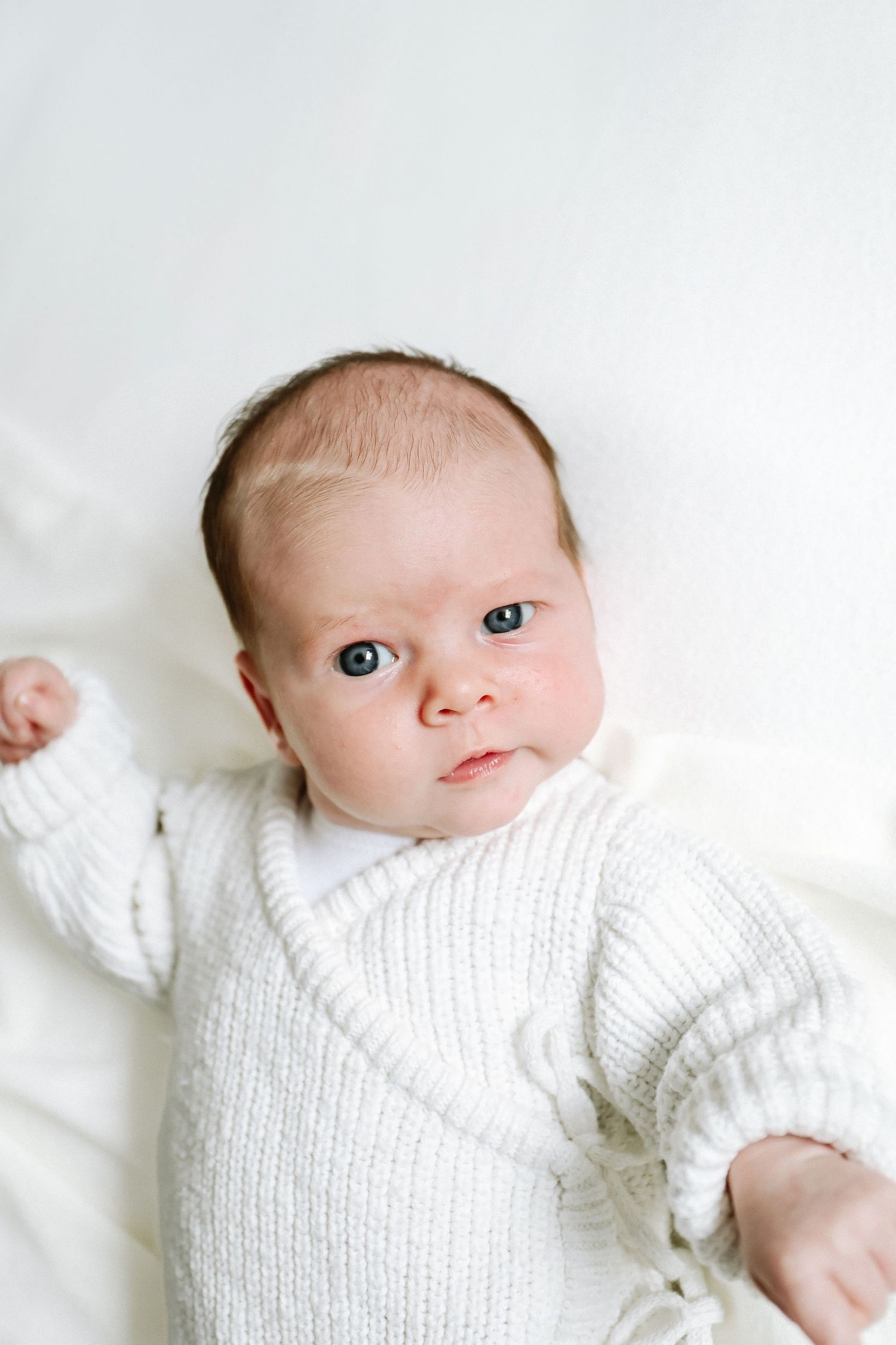 A tiny baby wearing a cozy white knit romper lays on a white surface while looking at the camera, during newborn photoshoot in Nottingham