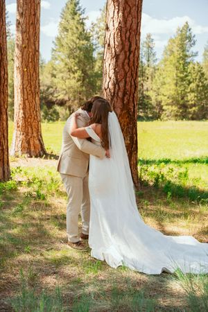 A couple embraces between tall pine trees during an outdoor wedding ceremony in a sunlit forest meadow.