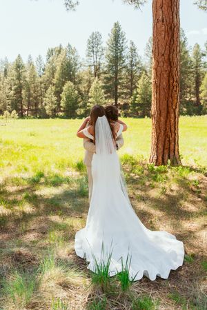 Long white wedding dress with flowing train photographed from behind in a sunlit forest meadow with tall pine trees.