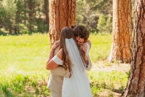 Couple shares a romantic embrace among tall pine trees in a sunlit forest meadow at golden hour.