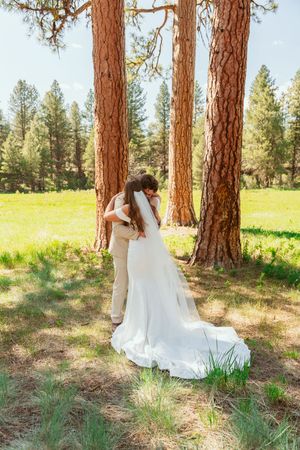 A romantic scene unfolds among tall pine trees in a meadow setting showing a white wedding dress train.