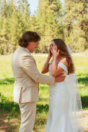 A couple in wedding attire shares their first look moment in a sun-dappled outdoor setting with pine trees in the background.