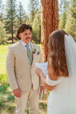 A wedding couple shares a sweet moment among tall pine trees in an outdoor ceremony setting.