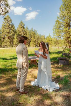Wedding couple dances together on a grassy meadow surrounded by evergreen trees on a sunny day.