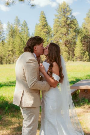 A tender moment is shared between a couple in light colored attire beneath towering pine trees.