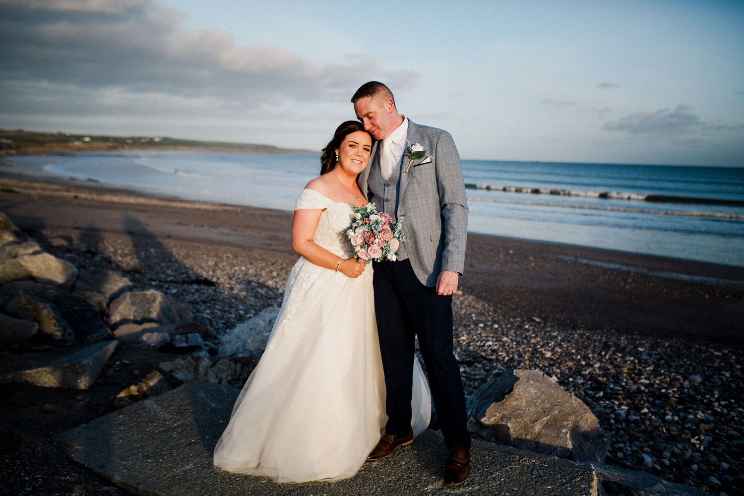 A romantic couple shares a tender moment on a rocky beach at sunset along the rugged coastline.