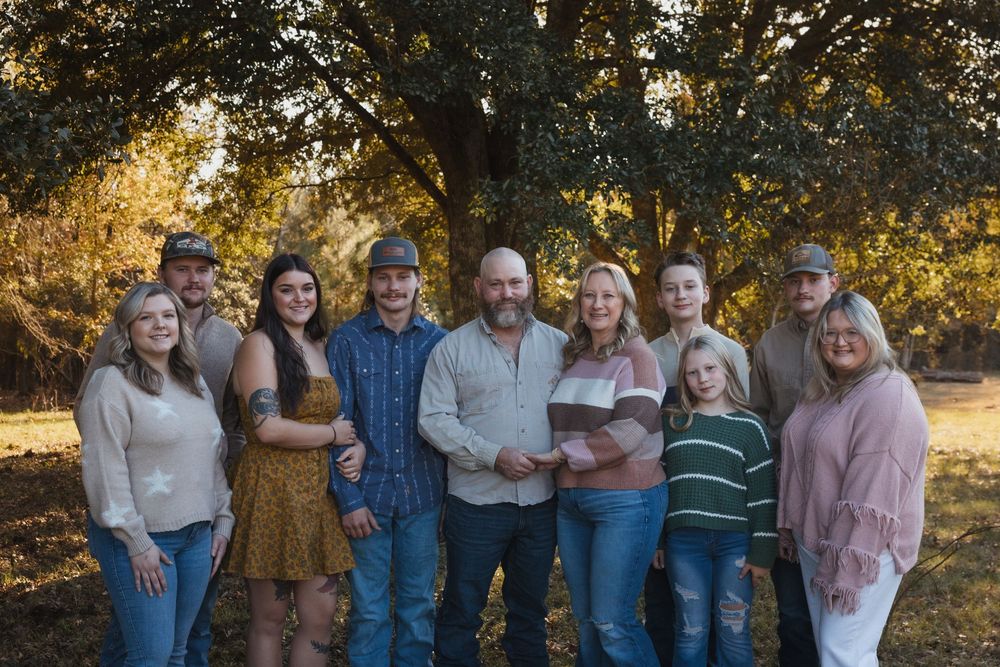Multi-generational family group poses together outdoors in casual attire during golden hour with trees in background.