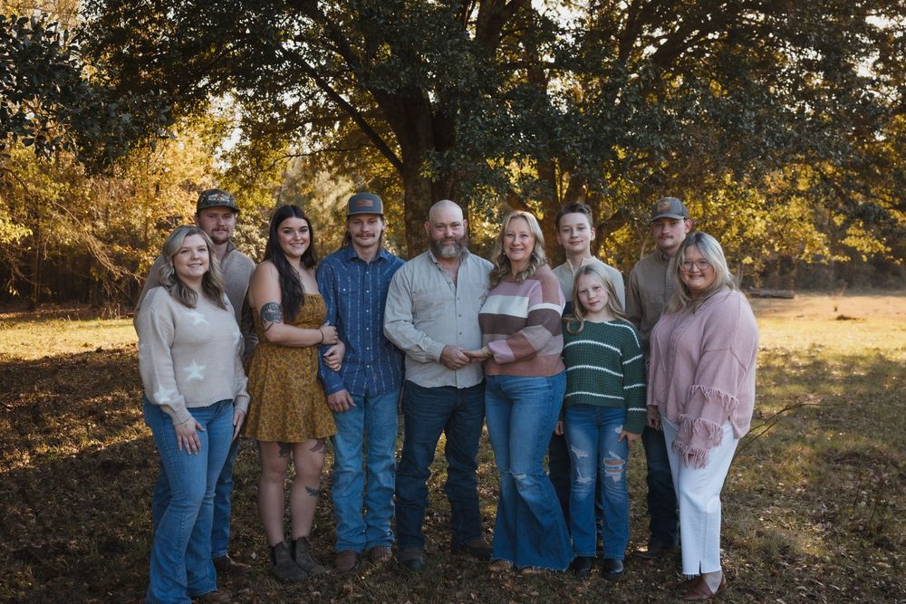 Multi-generational group poses together outdoors on a sunny autumn day with trees in the background.
