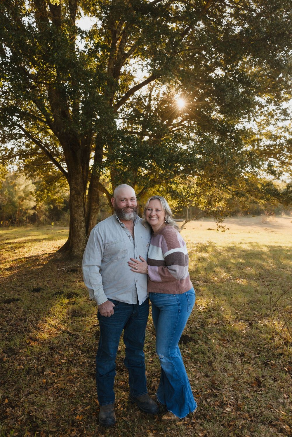 A couple embraces beneath a large tree during golden hour in an autumn field with fallen leaves.