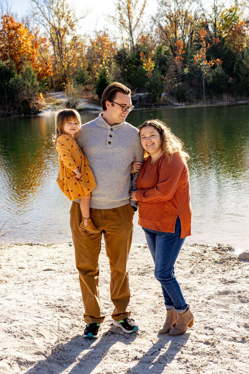 Family enjoying autumn weather together by a lake with fall foliage in warm earth tones.