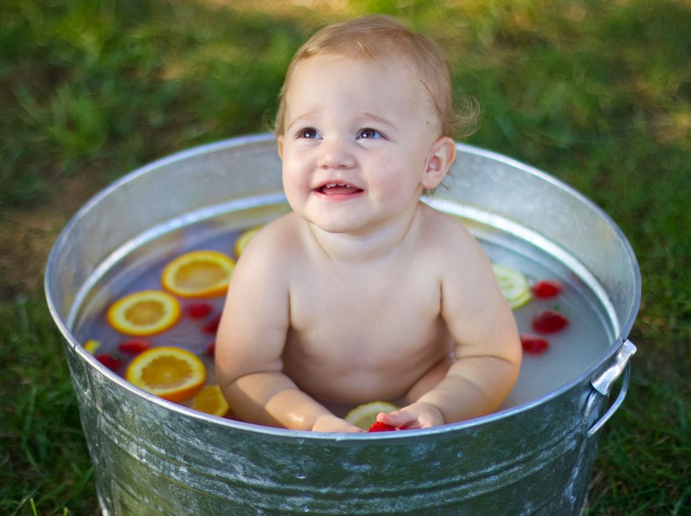 Baby sits in metal tub filled with citrus fruit slices on green grass.