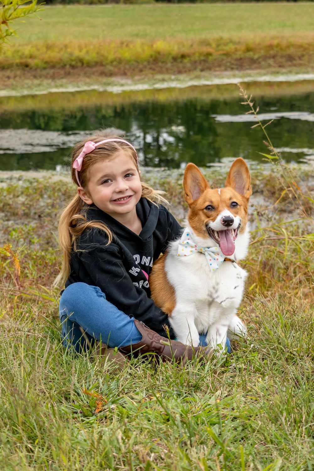 Child relaxing in grass with a corgi and white cat outdoors.