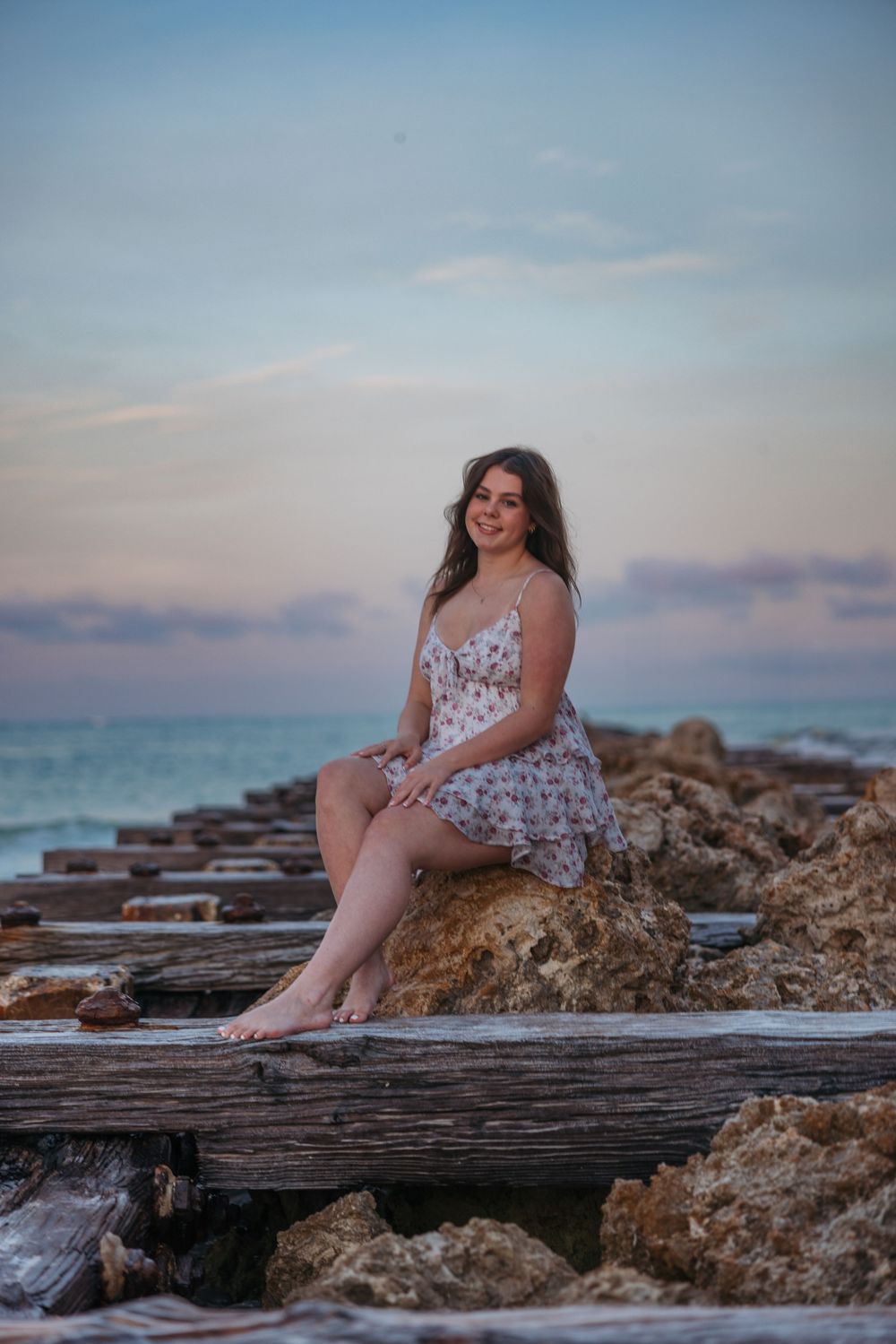 Young woman in floral sundress sits on wooden beams at seaside during sunset against turquoise ocean backdrop.