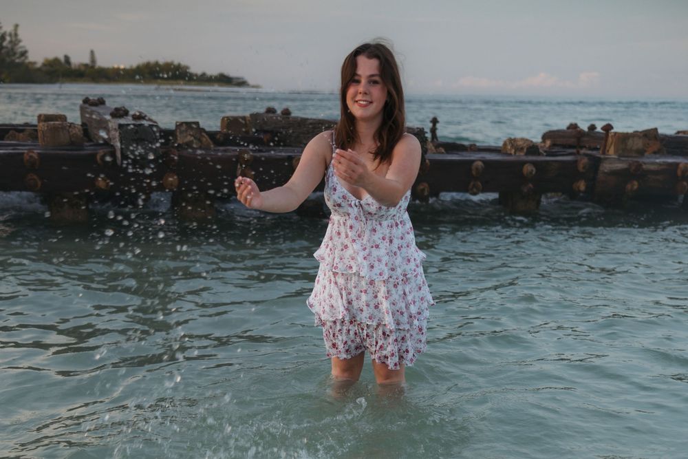 Playful moment splashing in ocean waters while wearing a white floral dress among rocks.