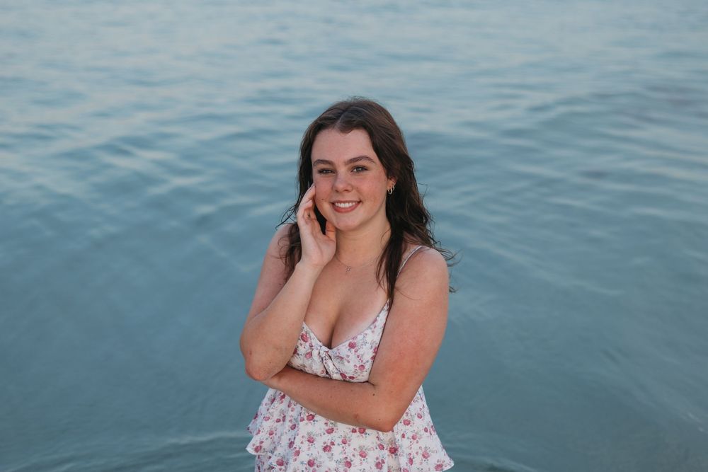 A figure in a white floral dress poses against a serene blue water backdrop.