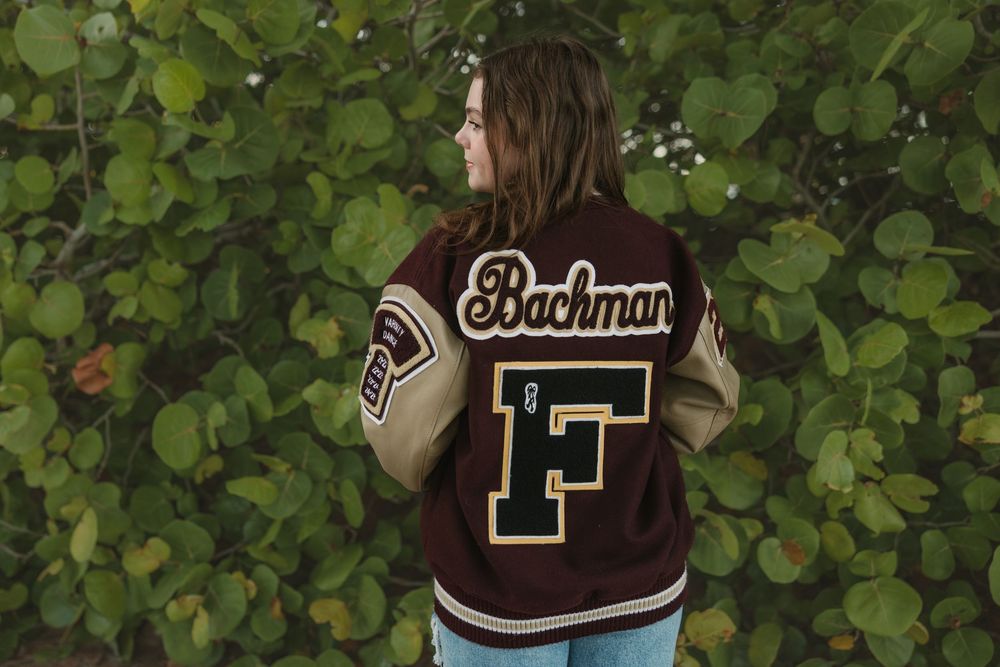 A varsity letterman jacket with the letter F is shown against green foliage.