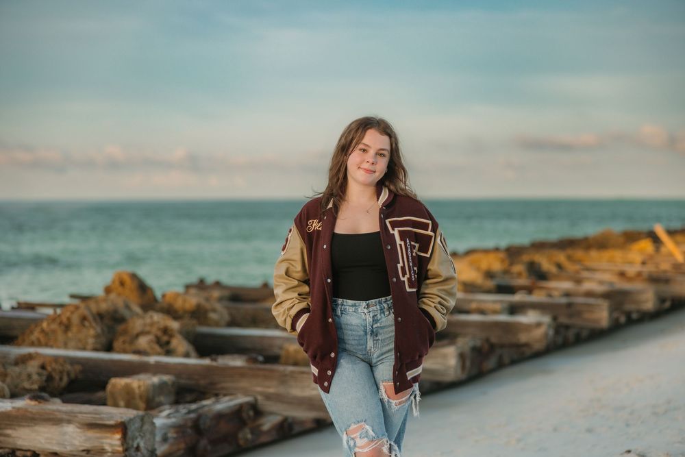 Figure in varsity jacket and jeans poses on oceanfront wood and rock jetty at dusk.