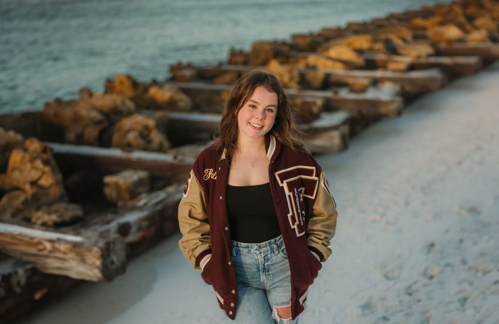 Someone in a varsity jacket and denim shorts poses along a rocky breakwater at the beach during golden hour.
