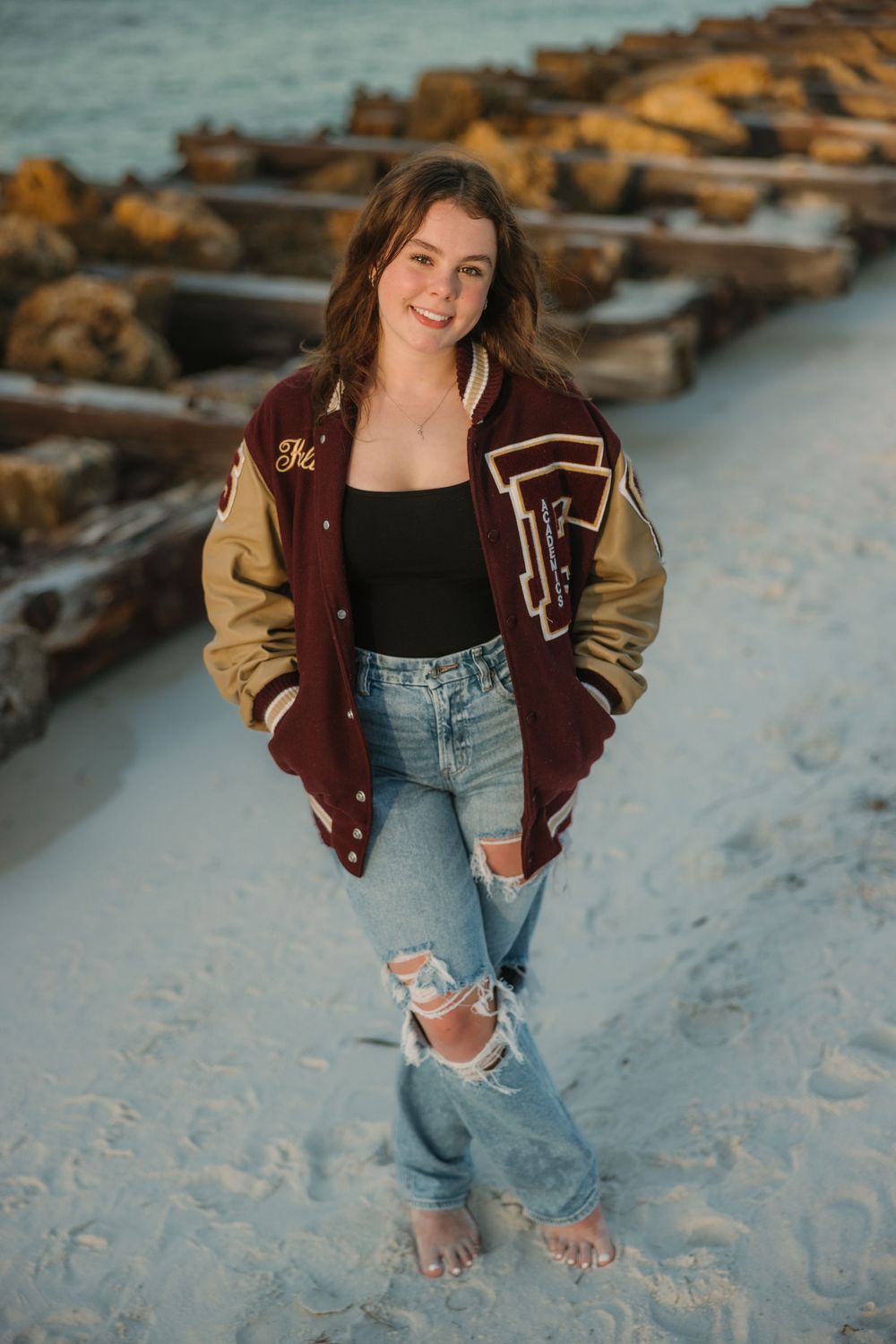 Casual beach style with varsity jacket and ripped jeans against coastal rockwork background.