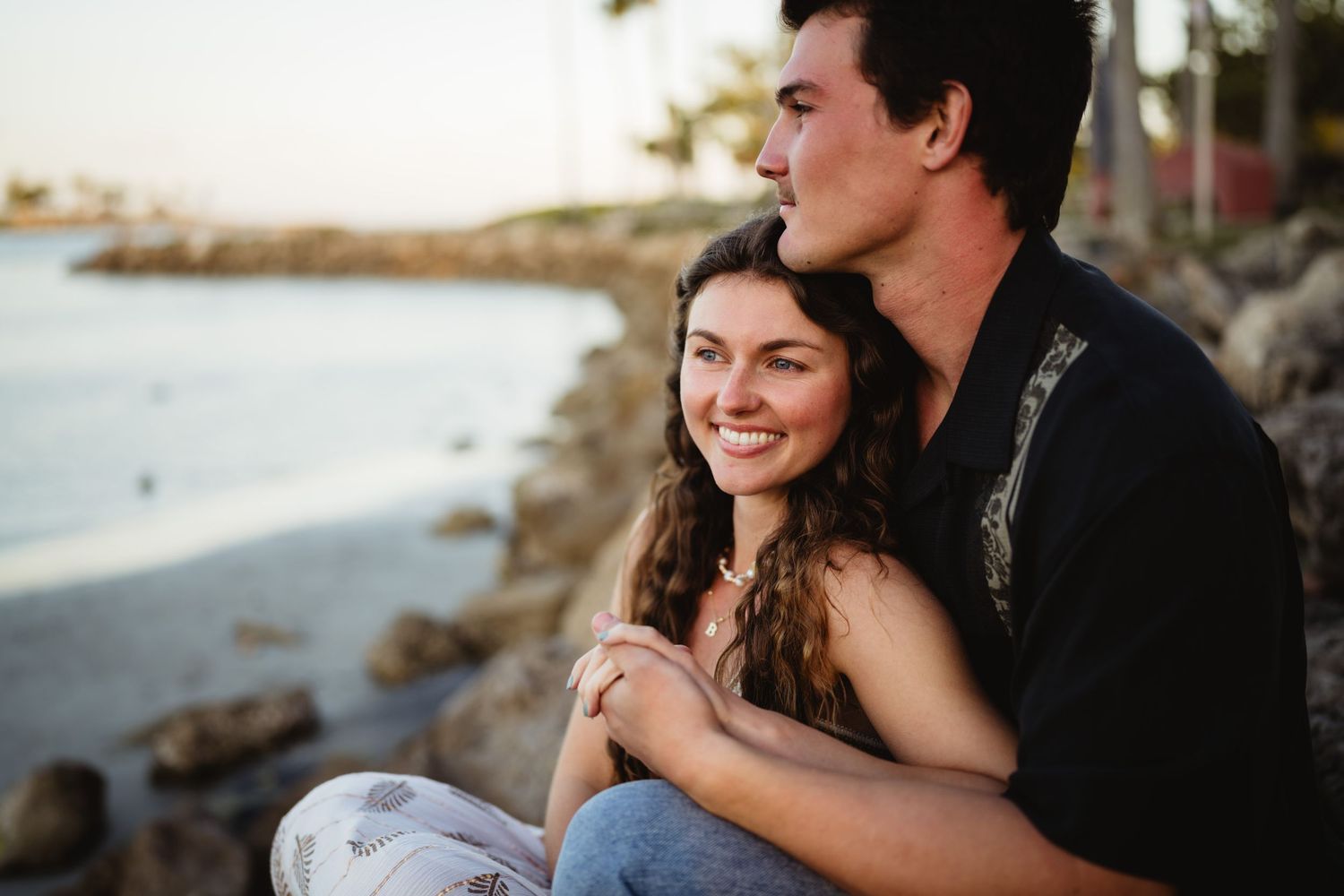 A couple sits together on coastal rocks at sunset, embracing while overlooking the ocean view.