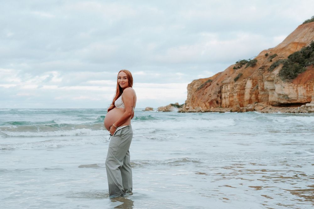 A maternity photo taken at the beach with rocky cliffs and ocean waves in the background.Altona Family Photographer Melbourne