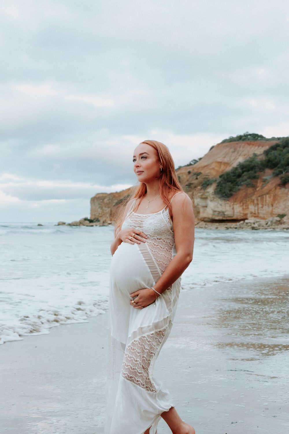 Expectant mother in white lace dress poses on rocky beach with cliffs in background during sunset.
Altona Family Photographer Melbourne