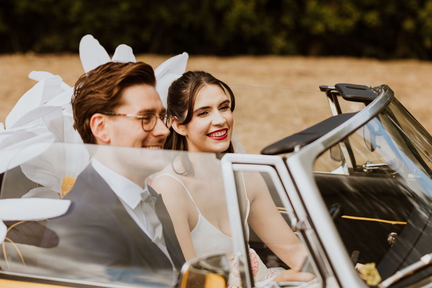A happy newlywed couple smiles while sitting in a vintage convertible car on their wedding day.