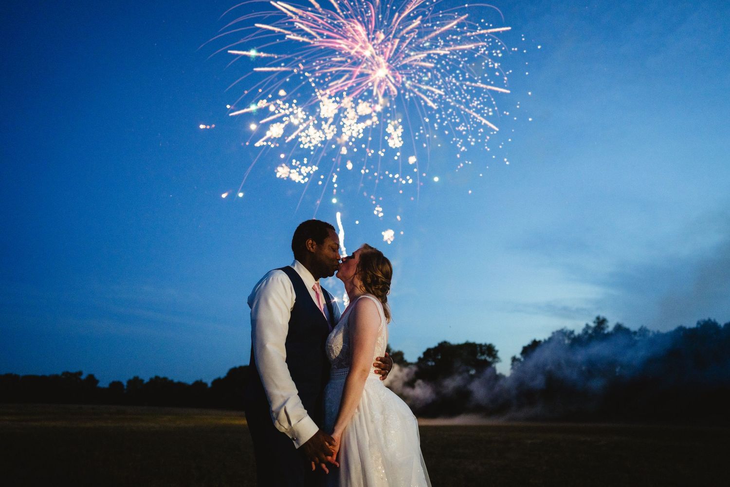 A couple in wedding attire shares a romantic moment under pink and white fireworks bursting in a twilight blue sky.