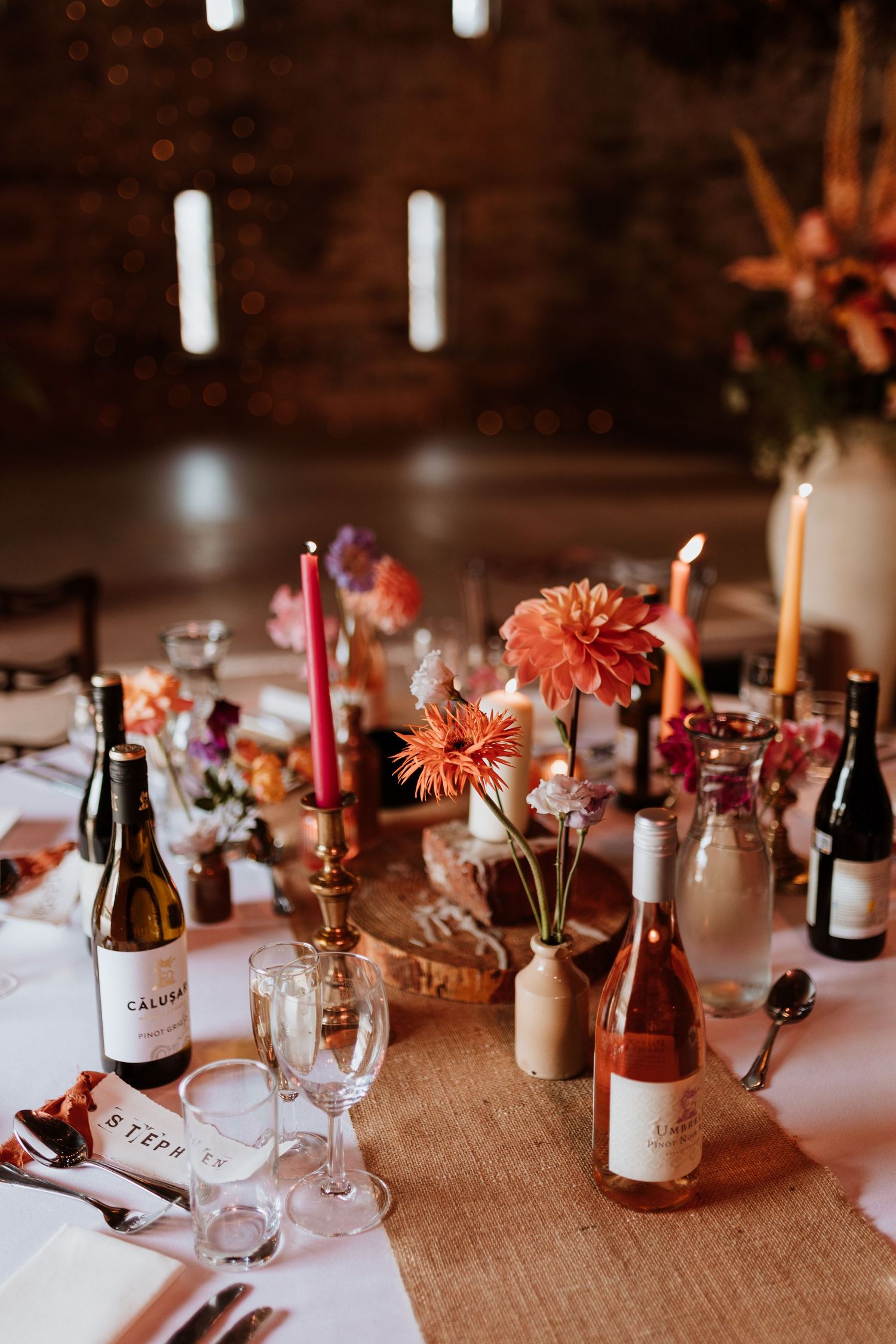 Rustic wedding table setting with coral dahlias, wine bottles, and burgundy candles on burlap tablecloth in warm lighting.