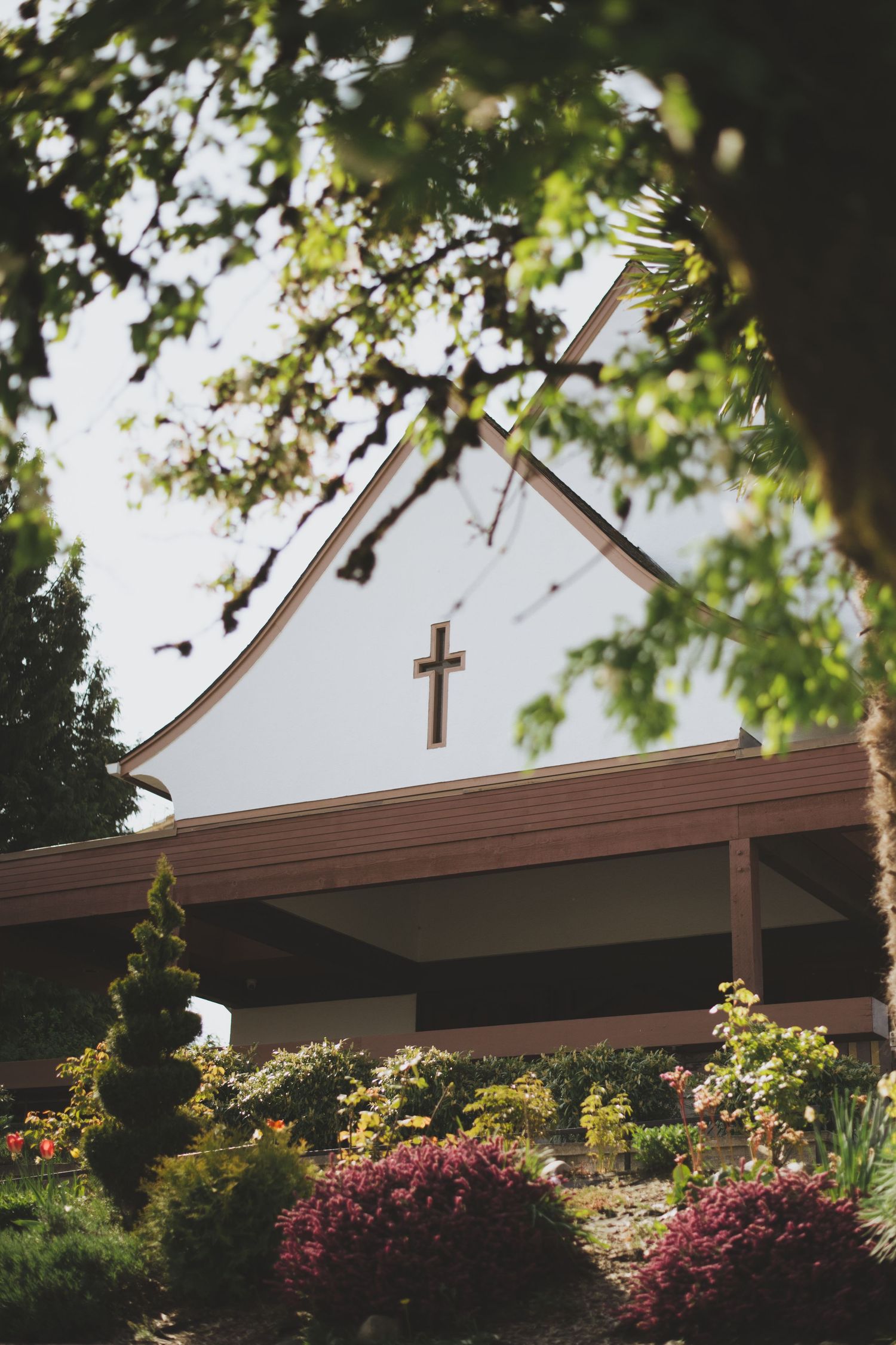 Cross on top of Holy Family Parish in Vancouver, BC, framed by green tree branches and pink flowering bushes.