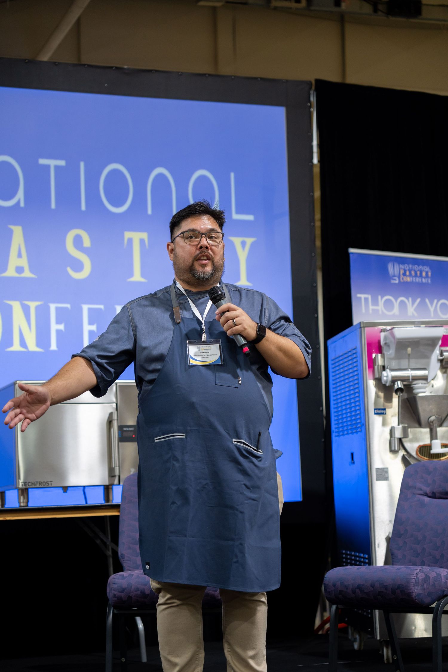 A presenter wearing an apron and glasses speaks into a microphone at a conference with stage equipment visible.