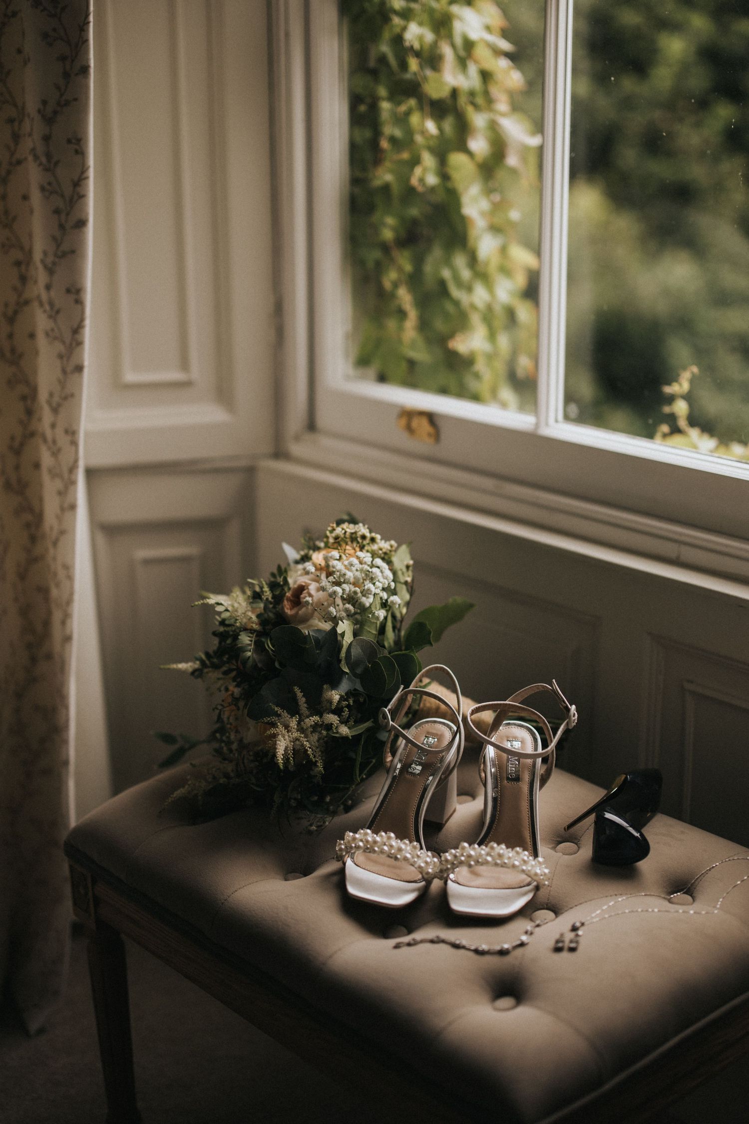 Elegant bridal shoes and bouquet arranged on tufted gray ottoman by window with natural light streaming in.