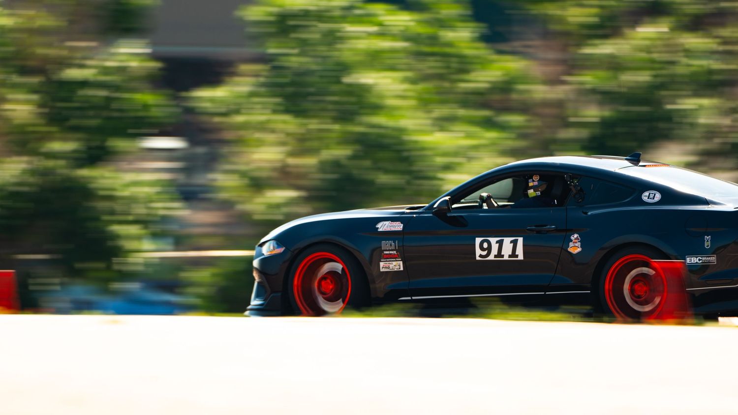 Black sports car with red wheels racing on a track with motion blur and green trees in background.
