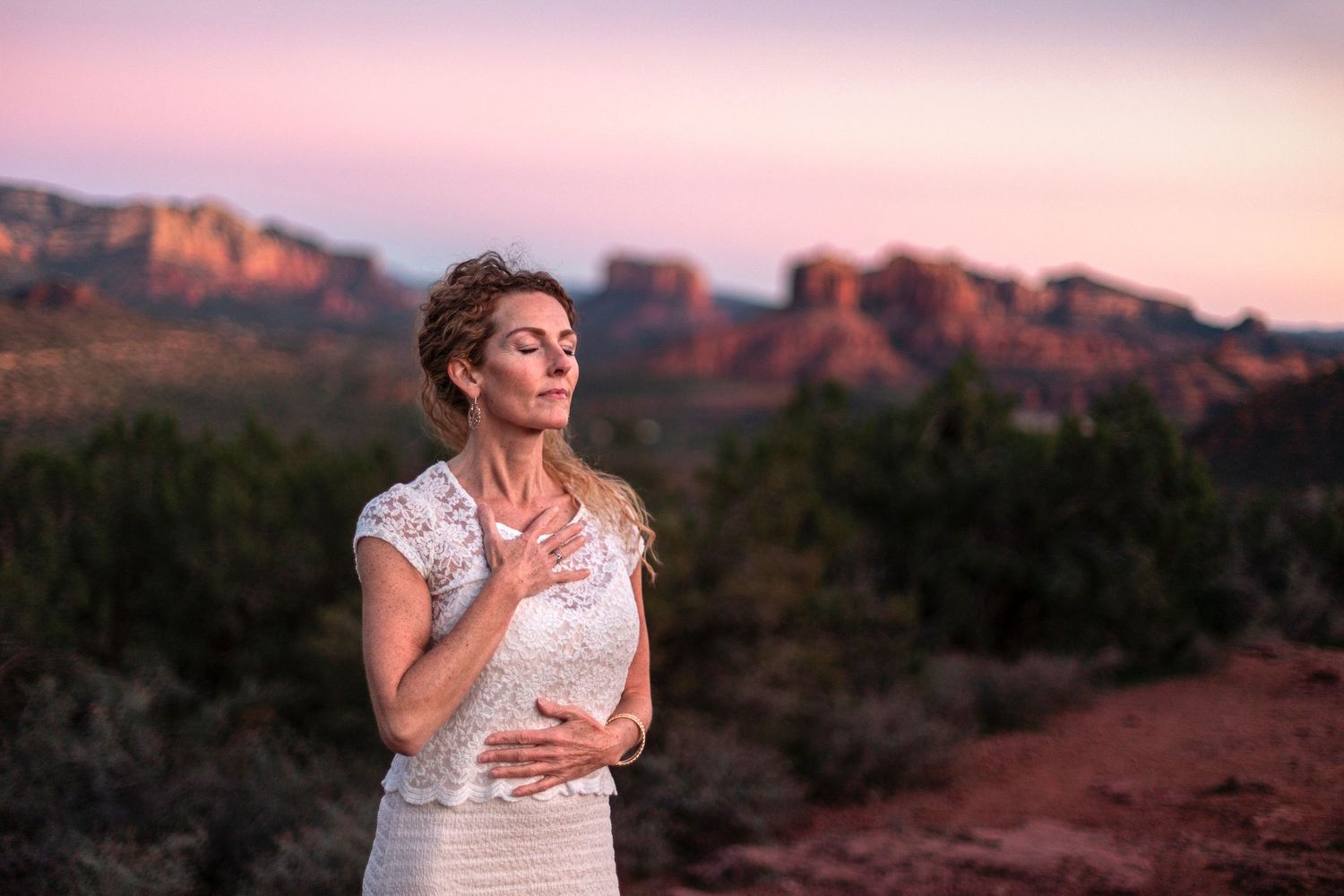 Serene woman in white lace dress with eyes closed at sunset in Sedona desert landscape.