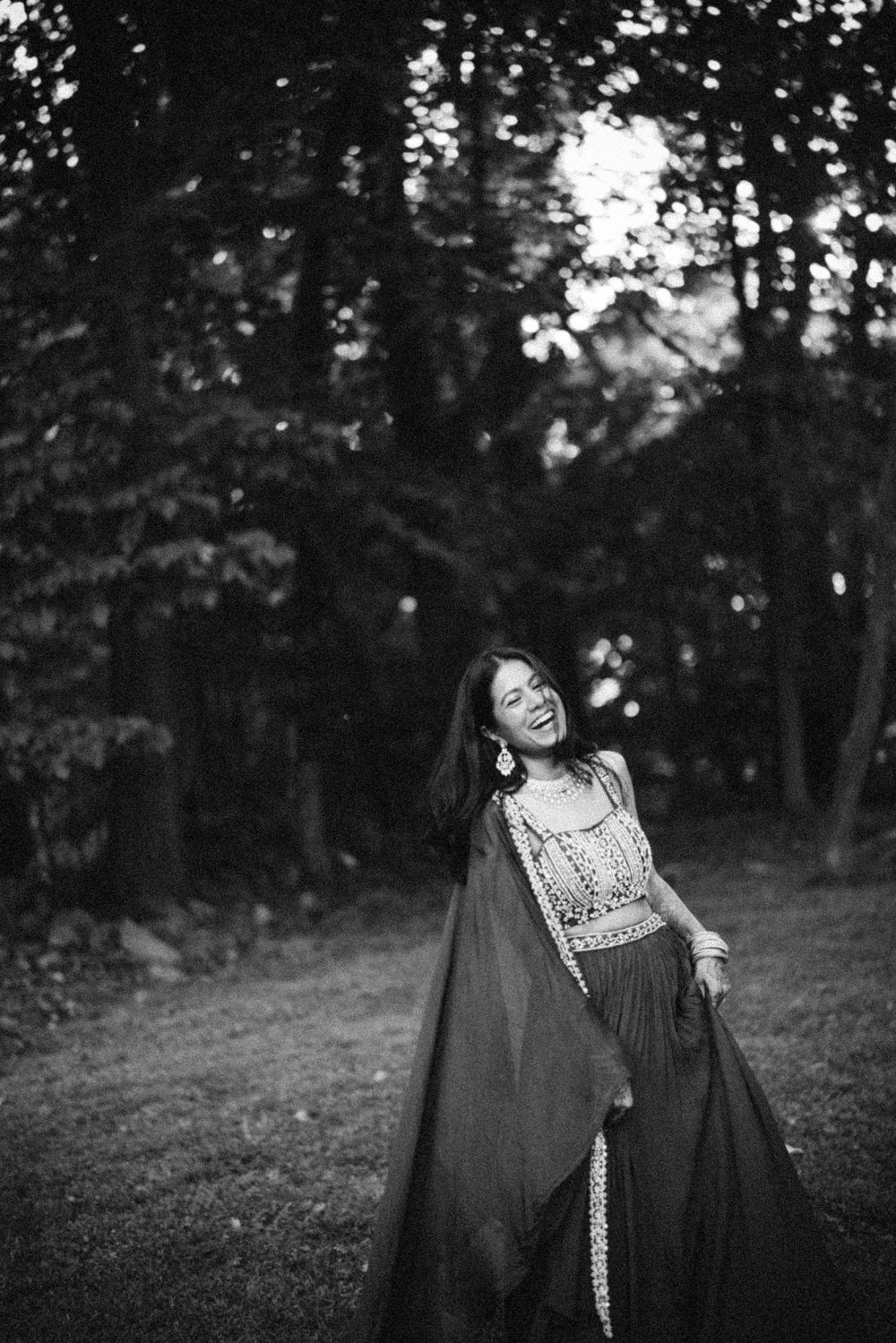 Black and white photo of a smiling woman in elegant skirt and embroidered top dancing outdoors in a forest.