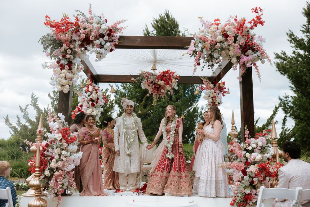 Indian wedding ceremony couple under floral mandap with red and white flowers, surrounded by family outdoors.