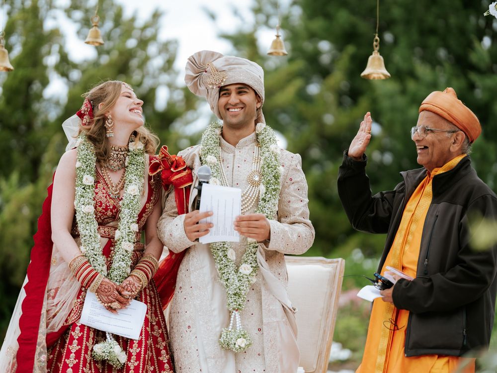 Indian wedding ceremony with bride in red sari, groom in sherwani, and officiant outdoors with floral garlands.