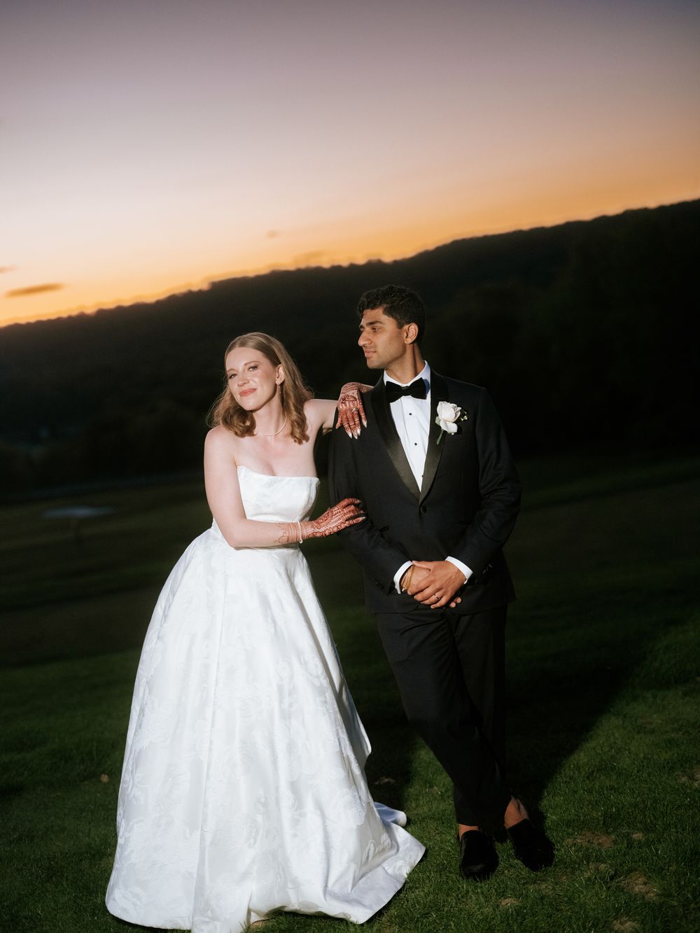 Bride in white gown and groom in black tuxedo pose together at sunset on a green hillside.