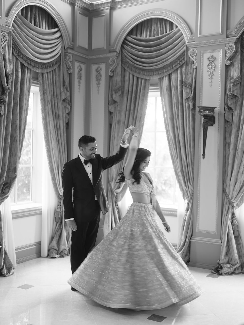 Couple sharing first dance in elegant ballroom, groom in tuxedo spinning bride in flowing gown, black and white photo.