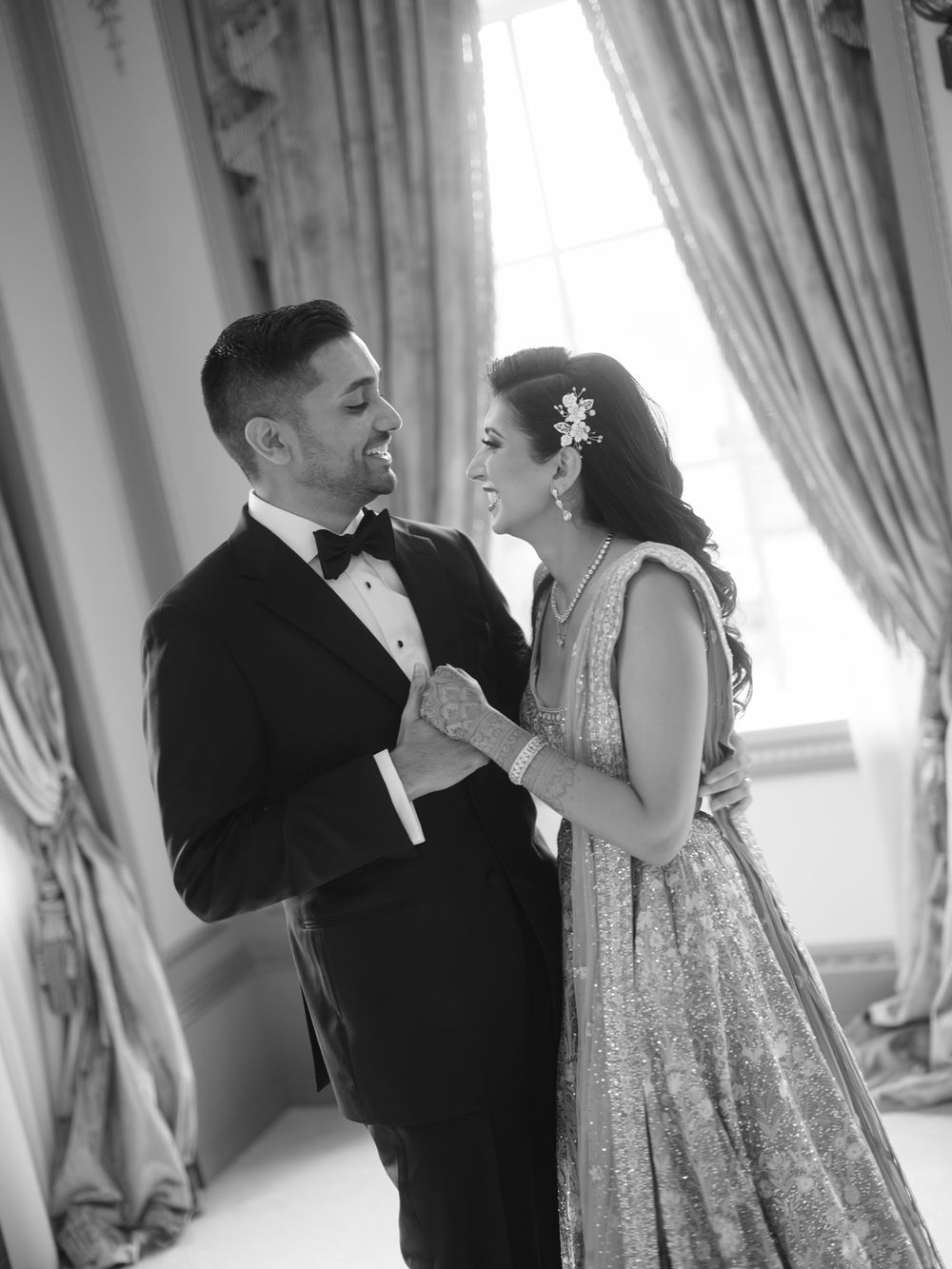 Black and white wedding photo of a couple in formal attire sharing a romantic moment by elegant draped curtains.