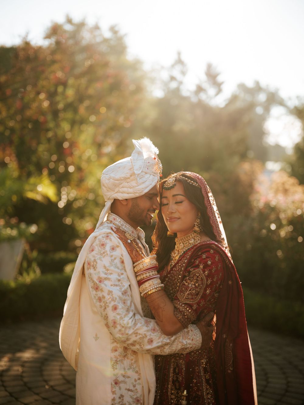 A couple in traditional South Asian wedding attire share an intimate moment in golden sunlight during their outdoor ceremony.