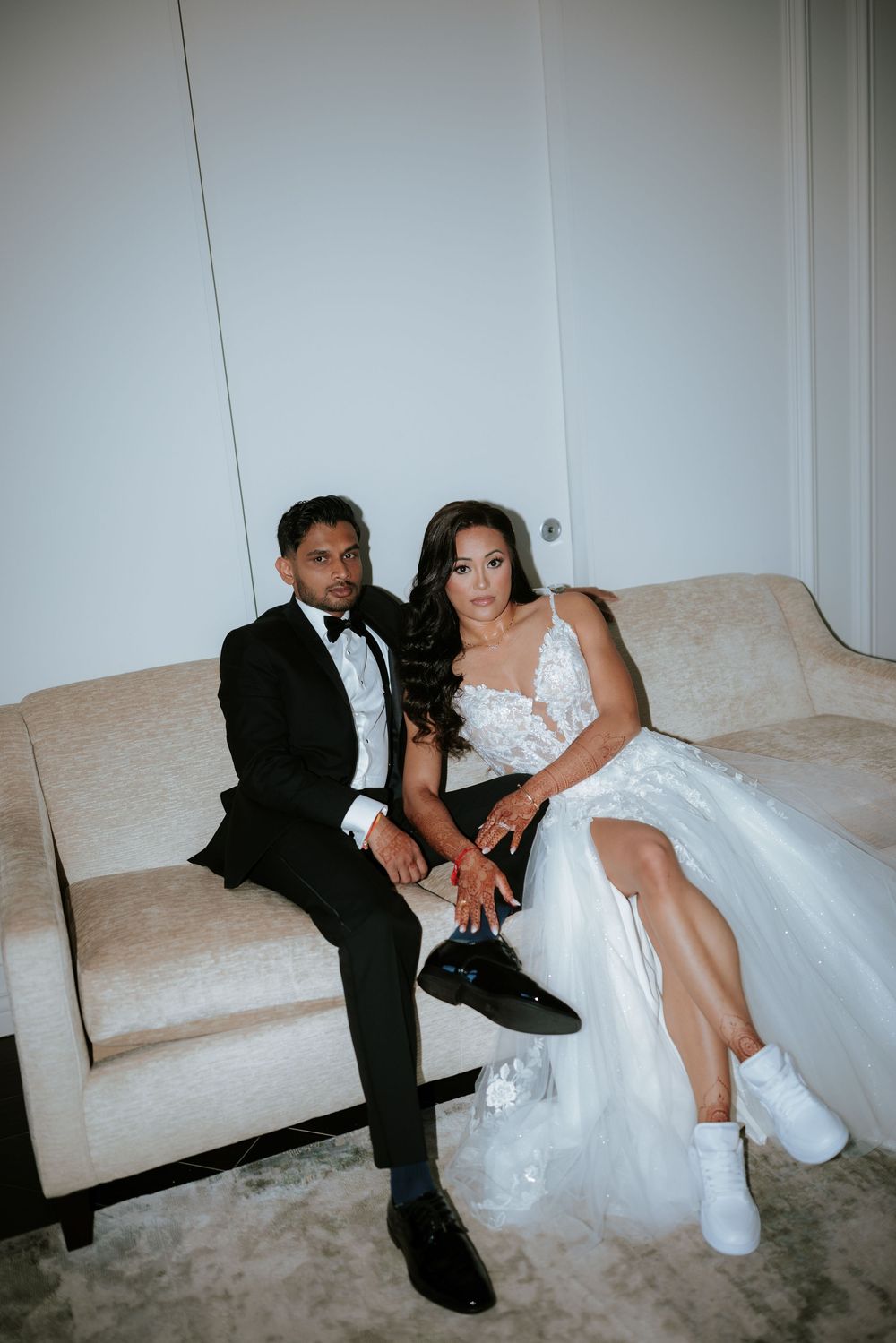 A couple in formal wedding attire sits casually on a beige couch wearing sneakers with their elegant outfits.
