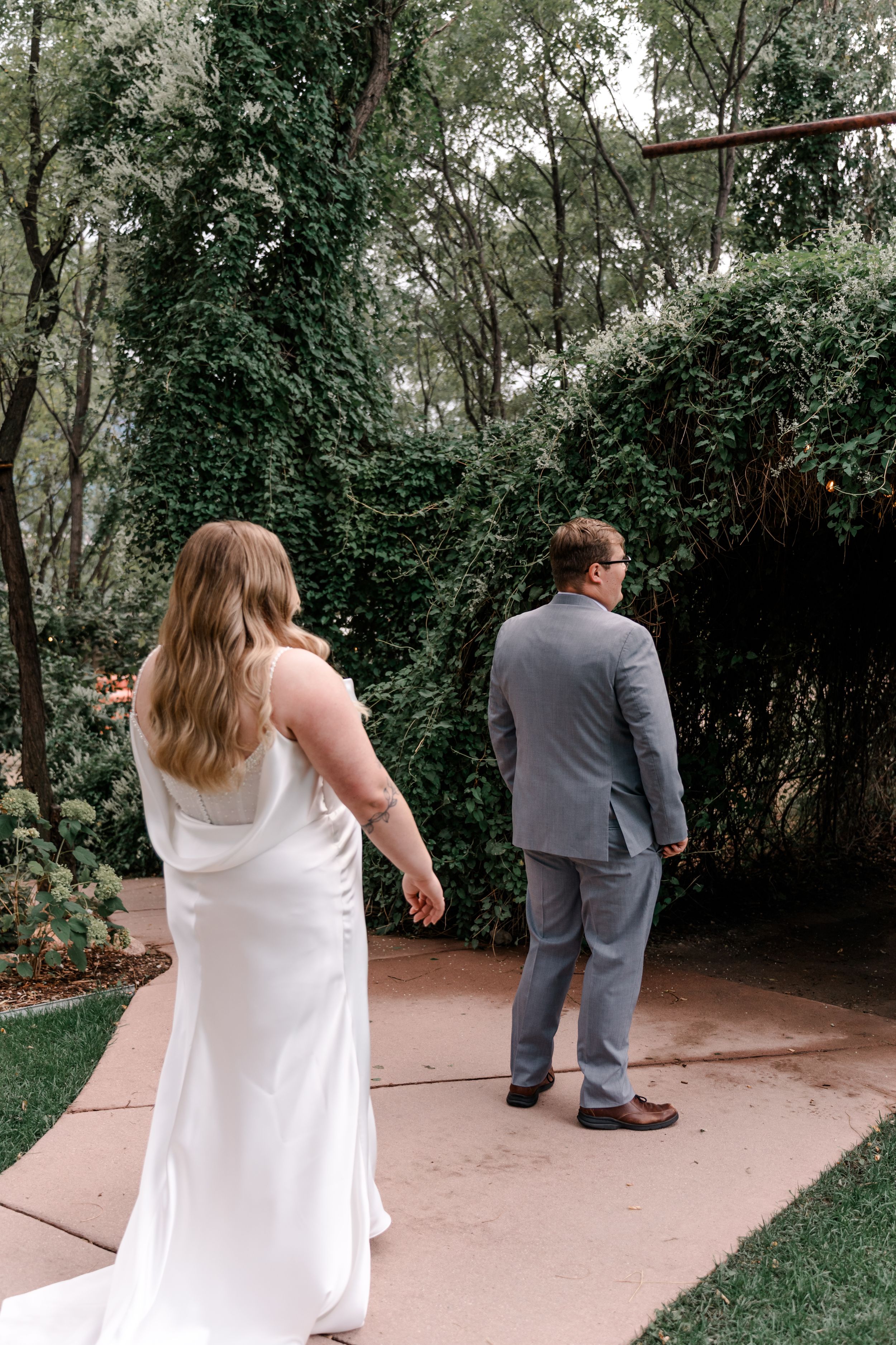 A couple in wedding attire walks along a garden path surrounded by lush greenery.