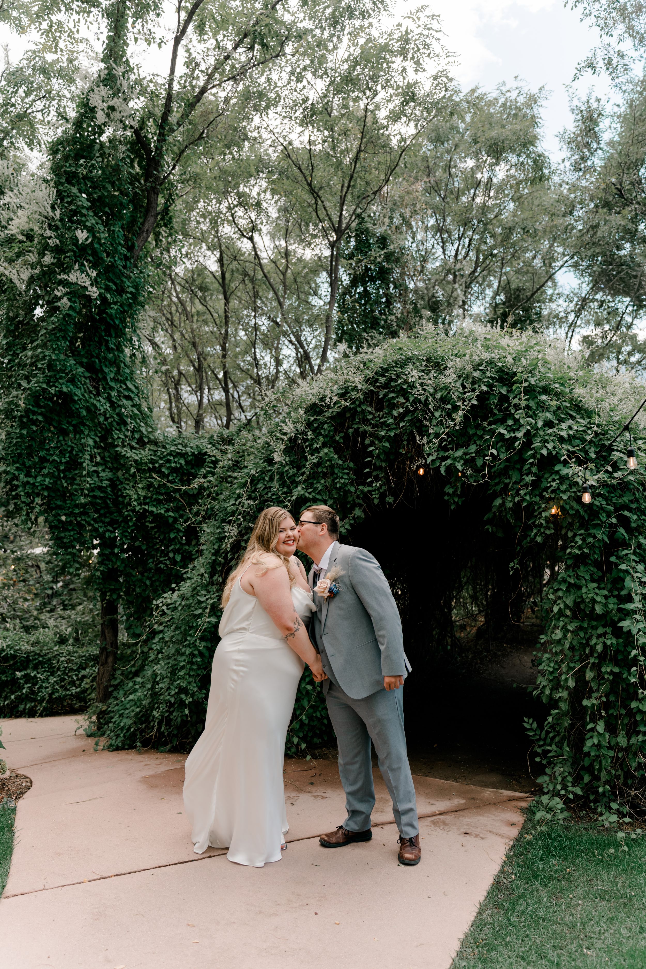 A romantic couple shares a tender kiss in an outdoor garden setting with lush greenery and trees in the background.