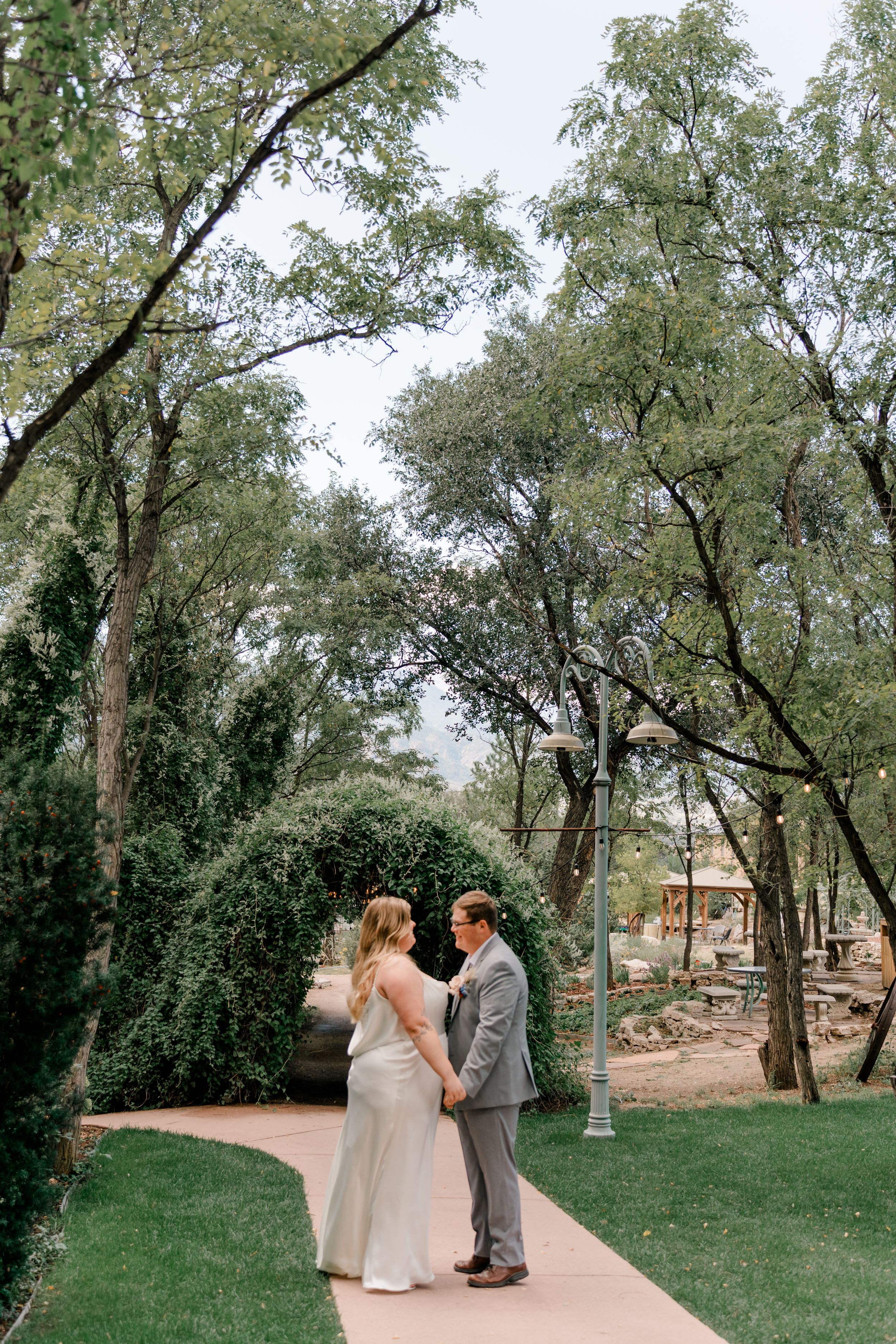 A couple embraces on a scenic outdoor pathway with trees and natural landscaping in the background.