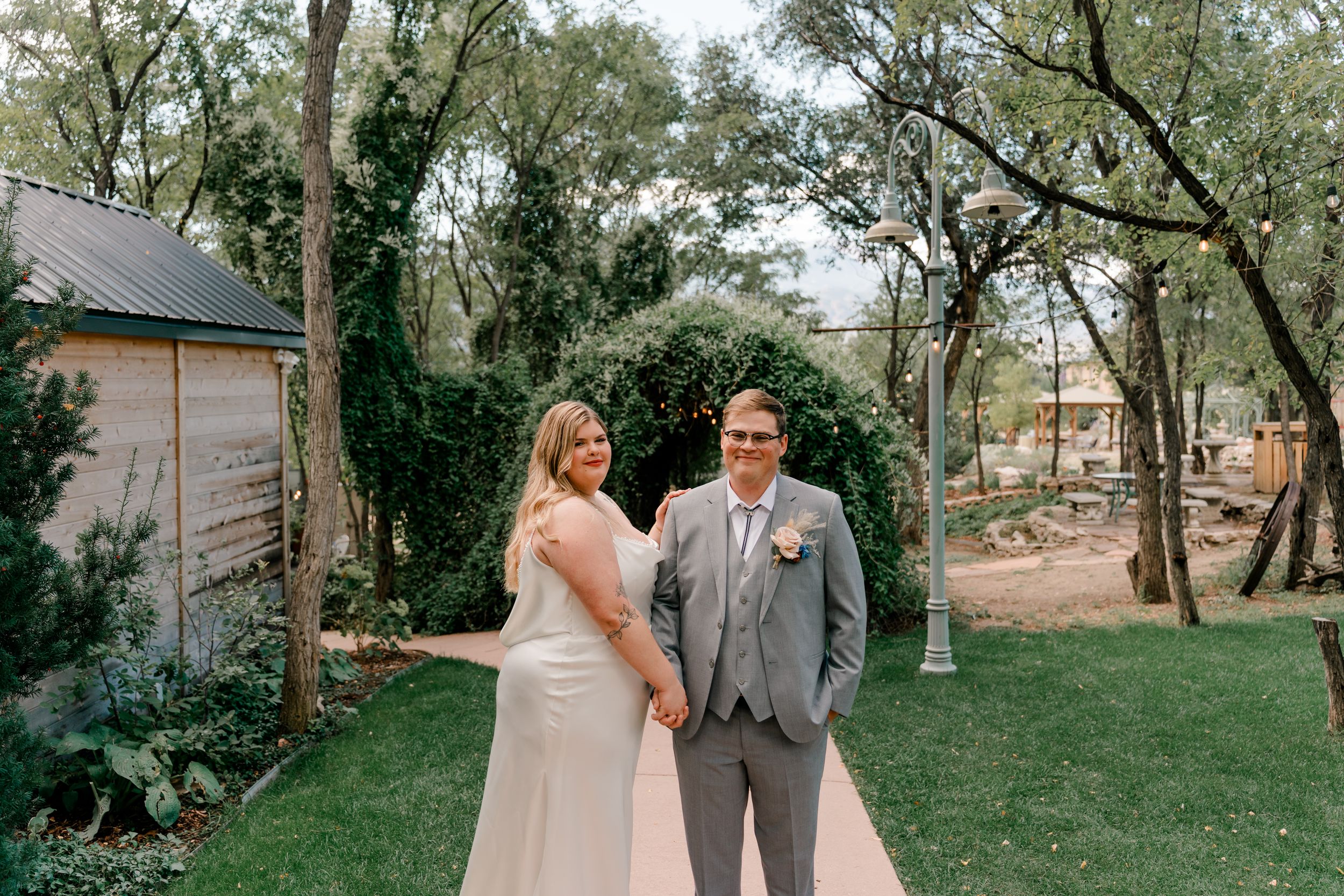 Wedding couple poses together on a grassy lawn with trees and rustic buildings in the background.