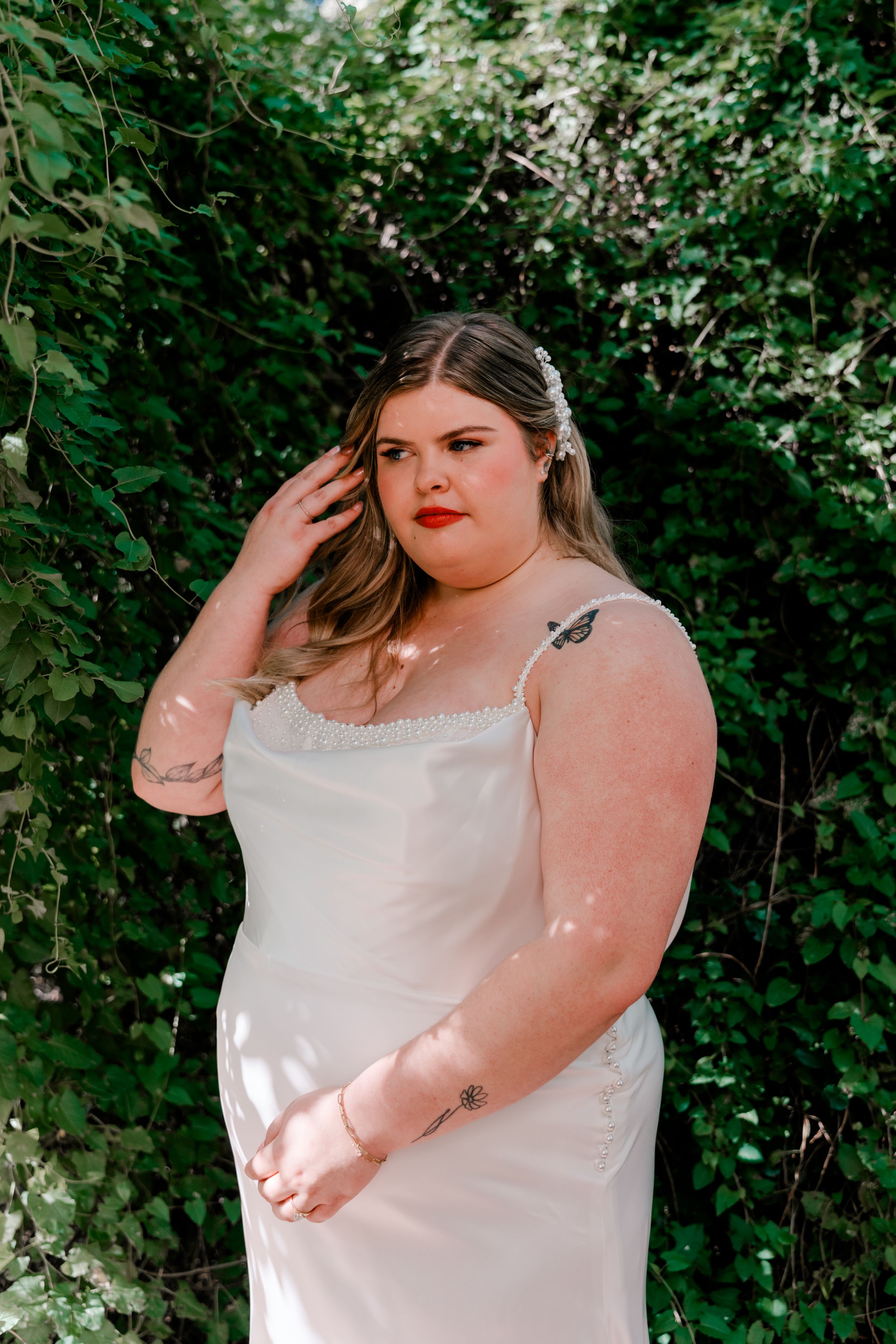 Bride in a white sleeveless dress poses against green foliage in natural lighting.