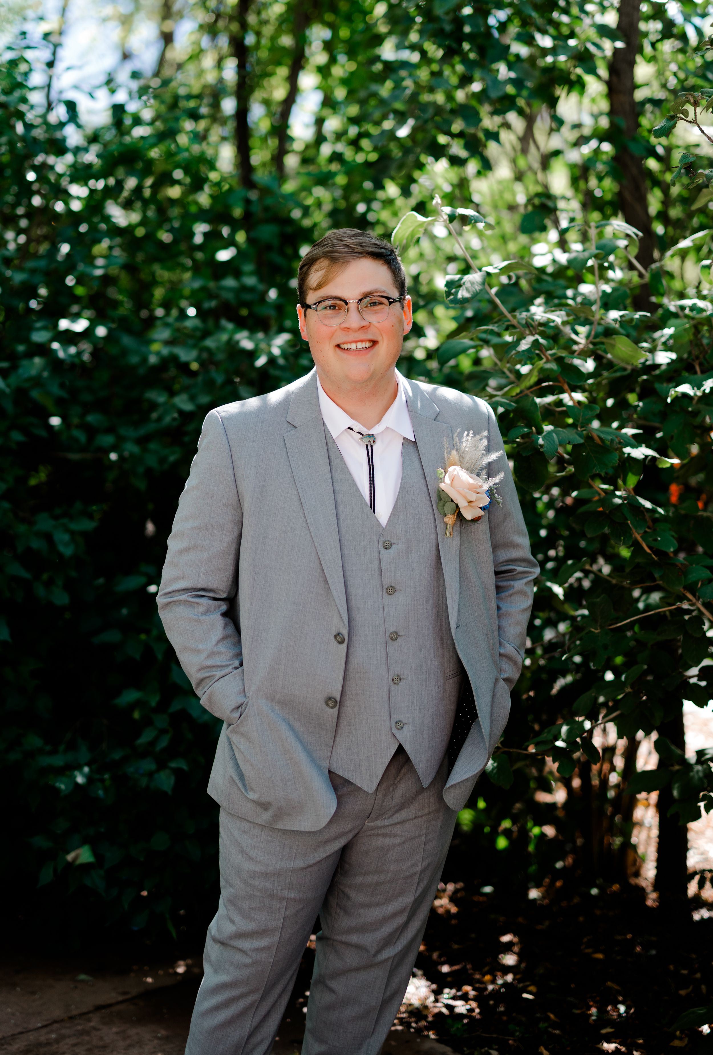Person in a light grey three-piece suit poses confidently in a garden setting.